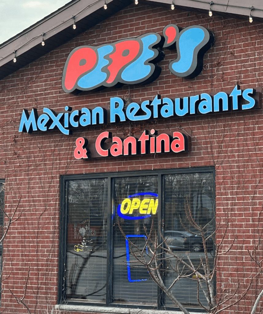 Exterior of Pepe’s Mexican Restaurants & Cantina with brick wall, colorful logo sign, and neon OPEN light in the window.